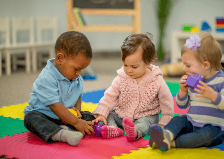 Three toddlers (two girls and one boy) playing on a colorful play mat divided into square blocks.