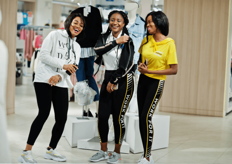 A lively scene featuring three young women wearing sporty outfits, smiling and posing in front of a mannequin in a clothing store. Their joyful expressions and casual style highlight a fun shopping experience.