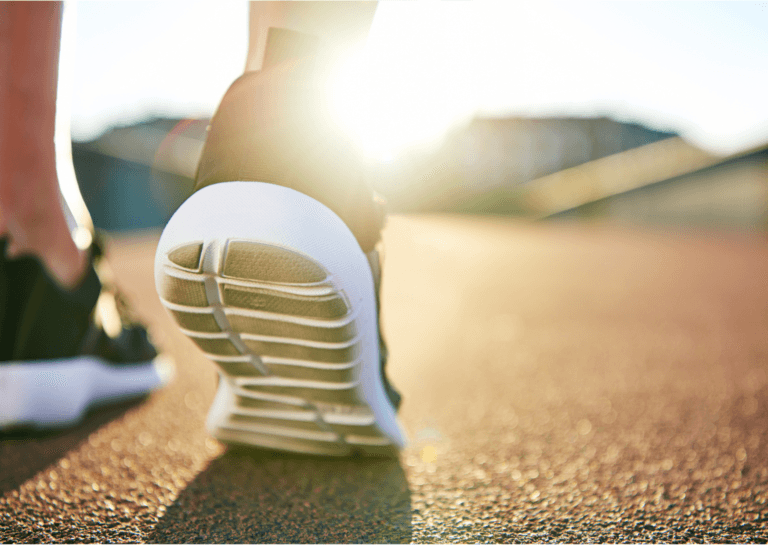 Close-up of training shoes worn by a person from behind, with the heel of one shoe slightly lifted, set against the backdrop of a sunset, filmed from the ground.