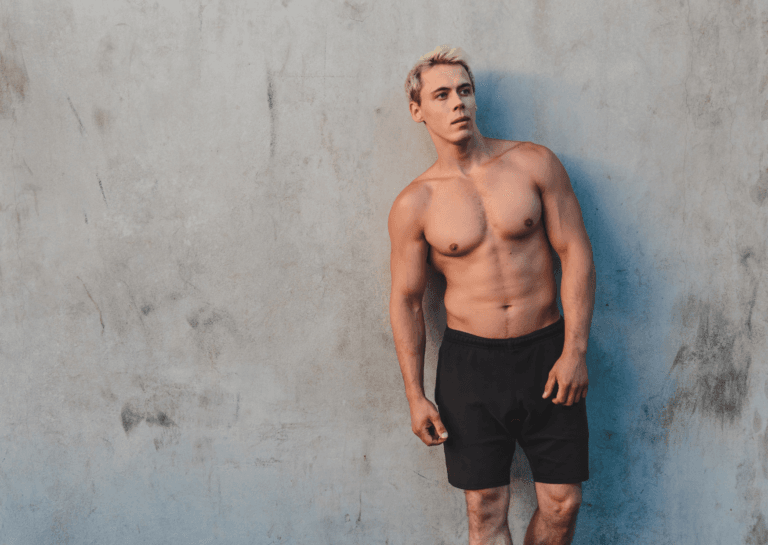 Muscular man with an exposed upper body wearing black shorts, looking away from the camera against a gray concrete wall backdrop.