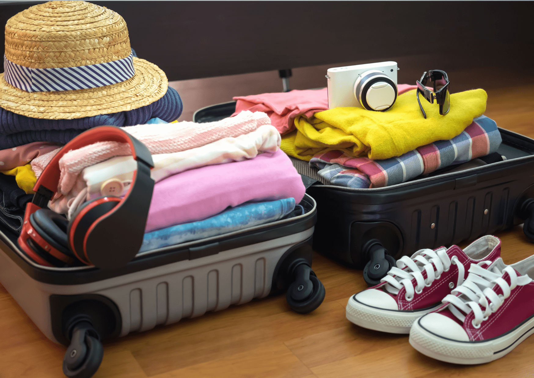 An open suitcase filled with a sun hat, clothing, camera, headphones, and All-Star shoes, all displayed on a light brown floor.