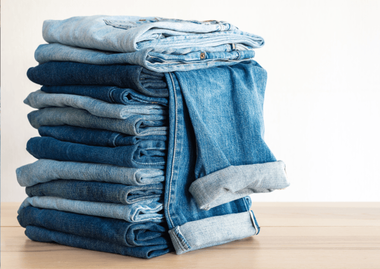 A neatly arranged stack of folded denim jeans displayed on a light brown table, set against a white background. The simple composition highlights the texture and colors of the jeans, emphasizing their casual, versatile style.