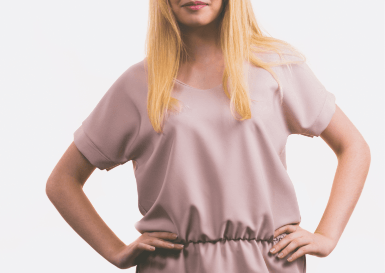 Blonde woman wearing a loose-fitting rose tunic, shown from the waist up with her hands on her hips, focusing on her midsection to her lips.