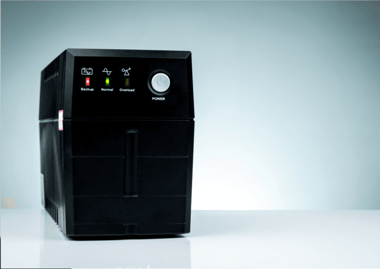 Black uninterruptible power supply (UPS) unit powered on, displaying red and green lights, positioned on a white desk.