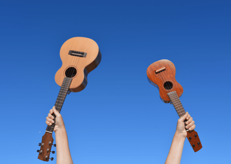 Two ukulele musical instruments held up against a blue sky, showing only two forearms with a light brown and a brown ukulele.