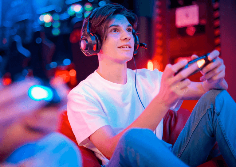 Young boy in a white shirt and jeans wearing black headphones, holding a game controller and smiling as he plays, set against a colorful, blurred background that resembles evening light.