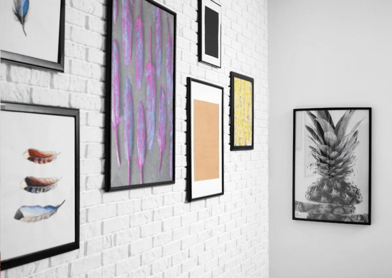 A corner of a room with a white brick wall on the left, decorated with various wall art, including three 'paintings' featuring feathers and three other pieces. The opposite wall is plain white, showcasing a painting of a pineapple.