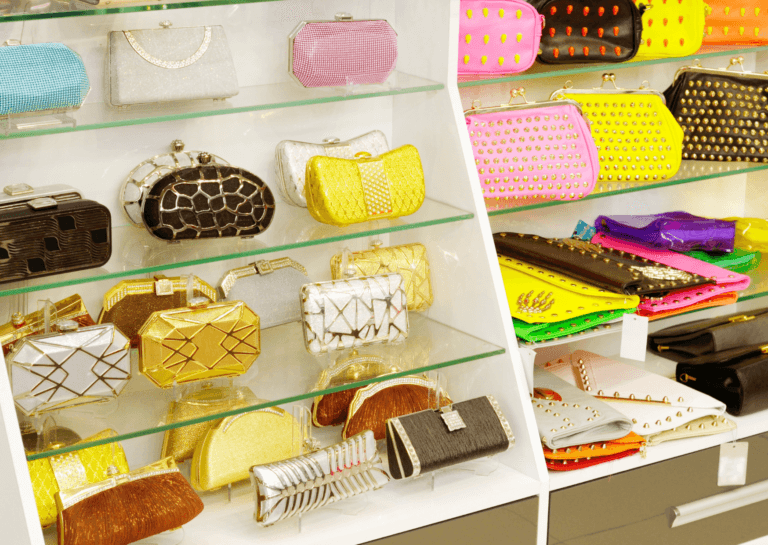 A vibrant assortment of wallets and clutches arranged on glass shelves against white walls. The collection features a range of colors, including brown, white, yellow, pink, blue, and purple, creating a cheerful and stylish showcase of accessories.