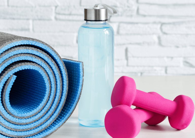 Close-up of two pink kettlebells stacked on top of each other on the right, with a rolled yoga mat on the left and a water bottle in the center, set against a white brick wall.