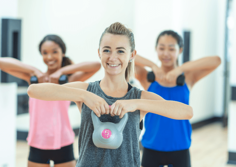 Three young women in a gym, with the two in the back holding dumbbells in both hands, while the woman in front is lifting a 5 kg kettlebell, all focused on their workout.