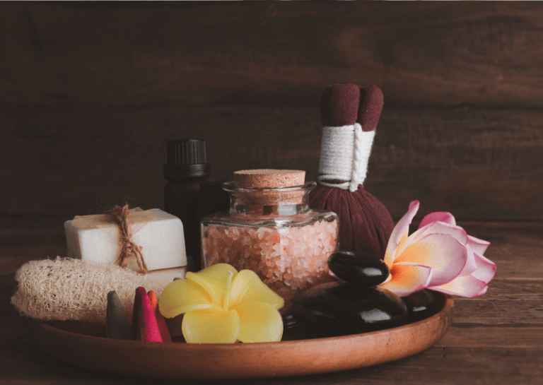 Round wooden tray featuring an array of wellness products, including bath salts, soaps, flowers, and massage stones, creating a relaxing atmosphere.