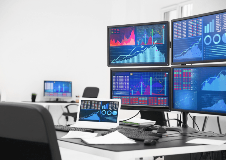 Workstation setup featuring four monitors—two side by side and two stacked above—along with an open laptop displaying graphs and market data. The setup is on a black desk with a white background.