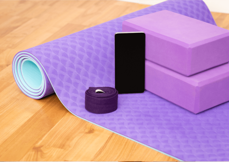 A half-rolled purple stretching mat features two stacked purple yoga blocks and a phone resting on top, alongside yoga straps.