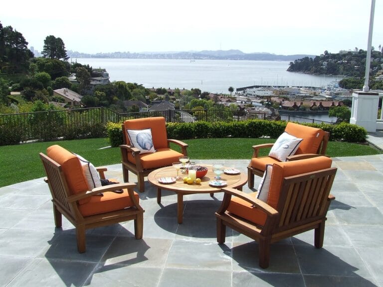 Teak patio furniture on a luxurious large terrace surrounded by low railings, with a beautiful bay and boats visible in the background.