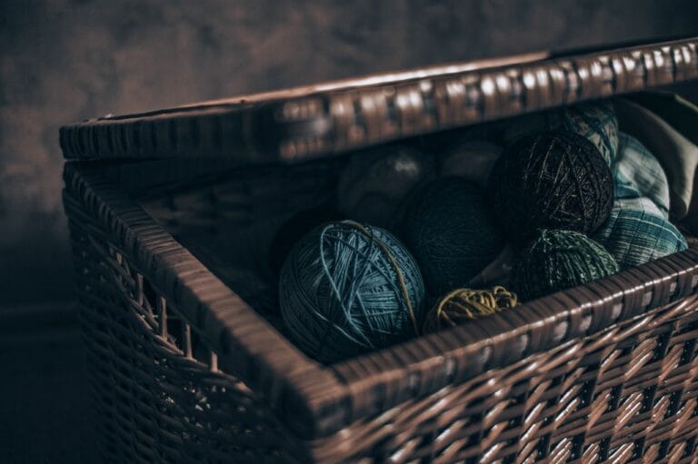 Brown woven storage box with a slightly open lid, revealing balls of yarn inside, set against a brown background.