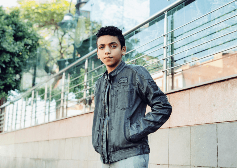 Young boy looking cool as he leans against a railing, facing the camera with his hands in the pockets of a stylish dark jacket, illuminated by daylight.