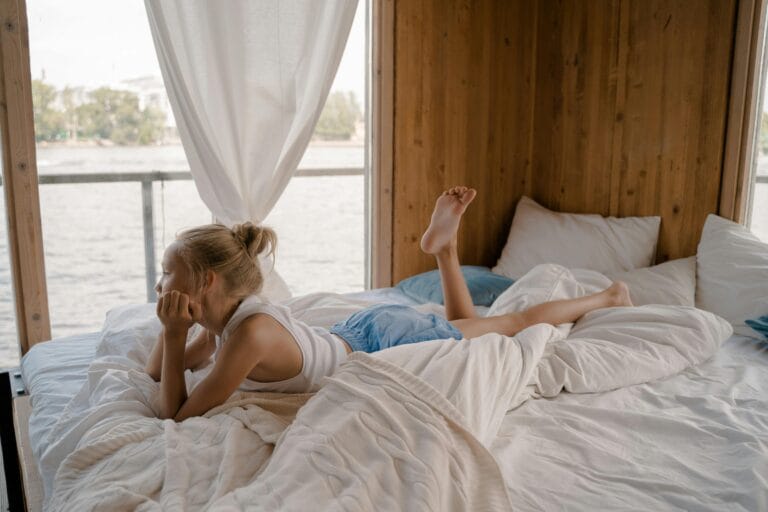 Blond girl lying on a bed and looking through a window at the water, as if she is in a bedroom of a houseboat.