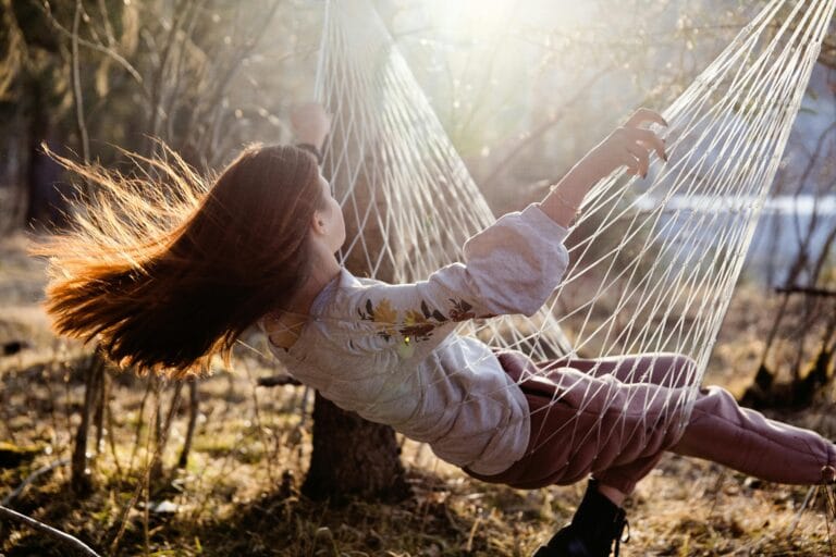 Girl with long red hair sitting in a hammock, swinging in nature with sunlight filtering through, her hair dancing in the breeze.