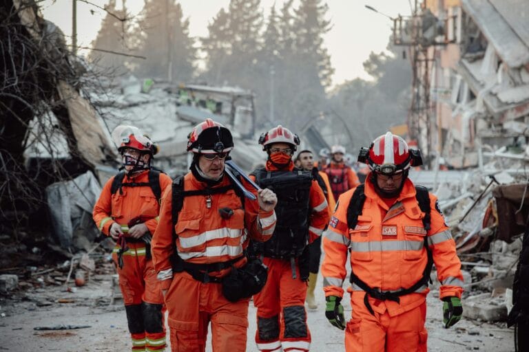 Group of paramedics wearing helmets and orange reflective suits walking through a demolished city, prepared for emergency response.