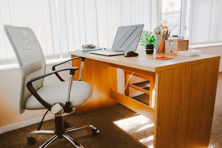 White rolling armchair beside an office desk in a bright and beautiful home office, featuring white hanging blinds that let in plenty of light.