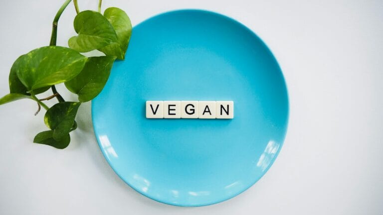 Scrabble tiles spelling the word 'vegan' arranged on a blue ceramic plate, accompanied by green leaves, all set against a white background.