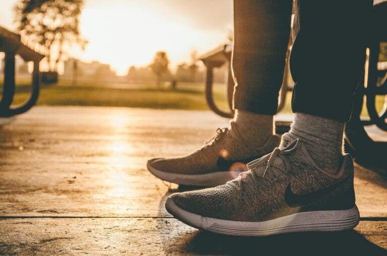 Person wearing sporty gray Nike sneakers and gray socks, sitting on a bench outside in black training pants, visible up to their calves, looking through the benches towards a city park bathed in the glow of the rising sun.