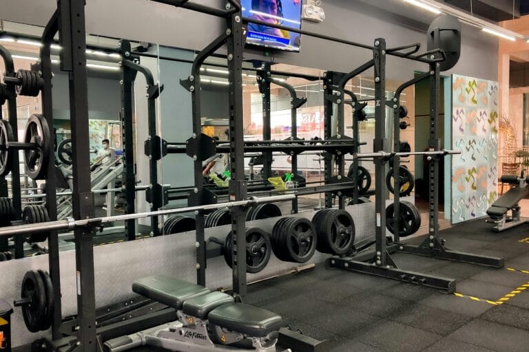 Exercise equipment set up on fitness mats in front of a mirror, featuring large machines like a squat rack, bench press, and heavy weights in a gym setting.