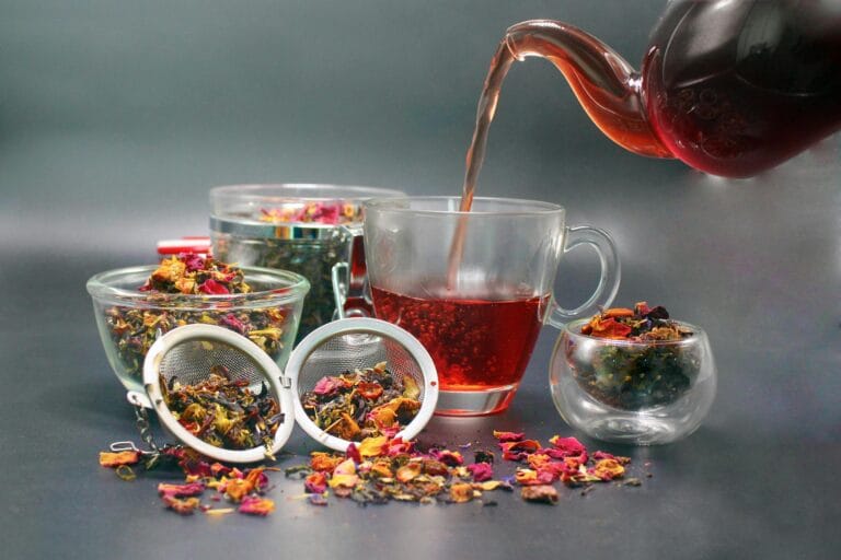 Fresh tea leaves in cups as tea is poured from a glass teapot into a transparent tea glass, surrounded by various containers of loose tea and an infuser ball for brewing.