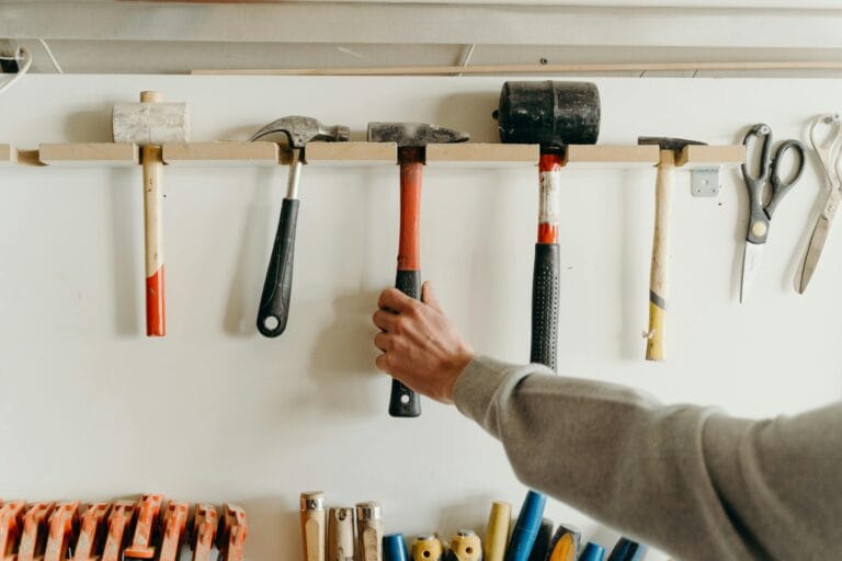 Person holding a hammer that is still hanging on a wall-mounted rack, featuring a variety of organized hammers.