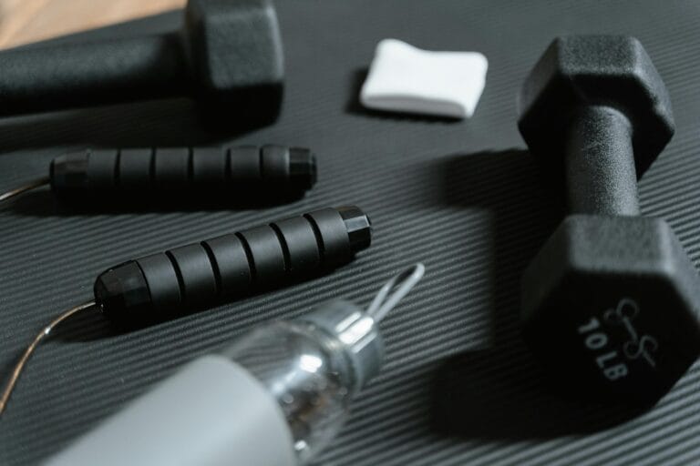 Close-up of dumbbells, a jump rope, a water bottle, and a sweatband on a black mat, creating an active fitness scene.