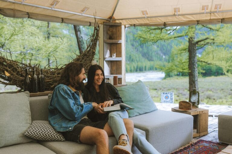 A couple sitting on a couch engaged in conversation under a large, cozy open shelter with a branch hammock, overlooking a river in the background.