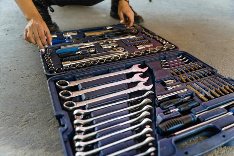 Open blue toolbox on the floor, filled with wrenches and ratchets, while a person crouches nearby, hands visible with white nail polish, reaching inside.